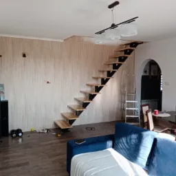 Nowoczesne schody z drewnianymi stopniami i metalową konstrukcją w jasnym wnętrzu domu, widoczne narzędzia na podłodze sugerują trwający montaż.