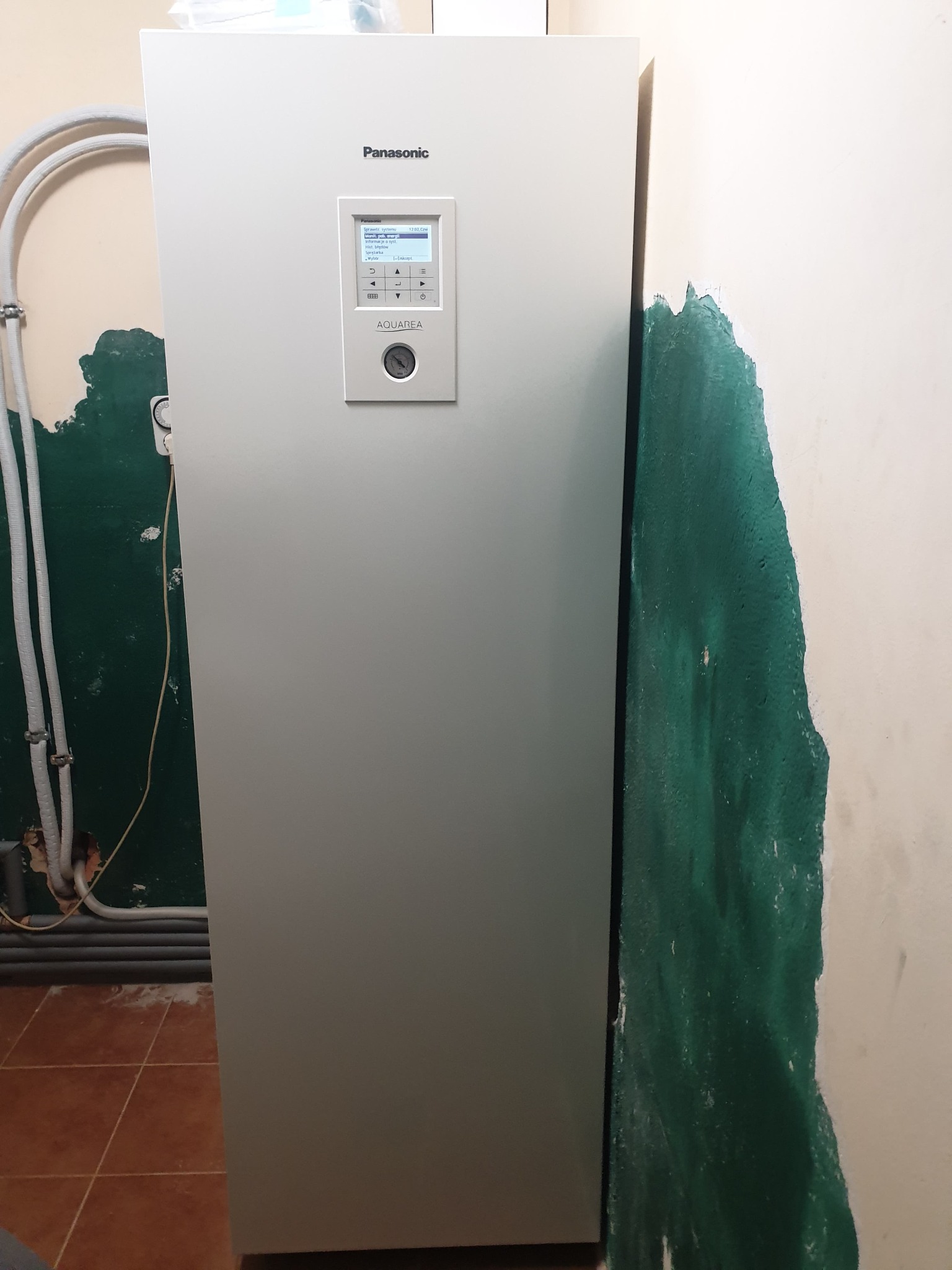Biała pompa ciepła Panasonic Aquarea zamontowana przy ścianie z widocznymi śladami po remoncie i instalacji rur.