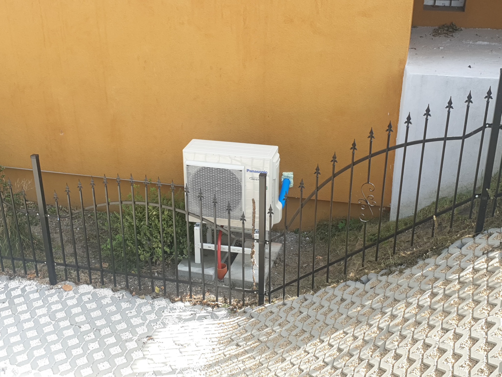 Zewnętrzna jednostka klimatyzacji Panasonic Inverter, umieszczona na betonowym fundamencie za czarnym, metalowym ogrodzeniem, na tle pomarańczowej ściany budynku.