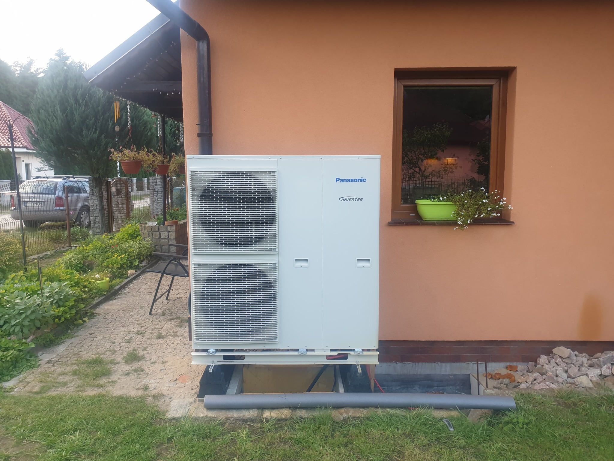 Zewnętrzna jednostka klimatyzacji Panasonic Inverter zamontowana na fundamencie przy ścianie domu z oknem, widoczne przewody i fragment trawnika.