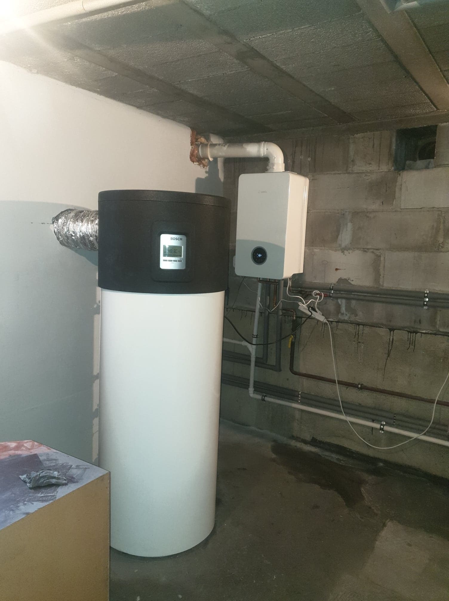 Biała pompa ciepła Bosch z czarną górną częścią i wyświetlaczem, podłączona do rury wentylacyjnej, obok wiszący na ścianie biały piec gazowy w surowym pomieszczeniu z betonowymi ścianami...