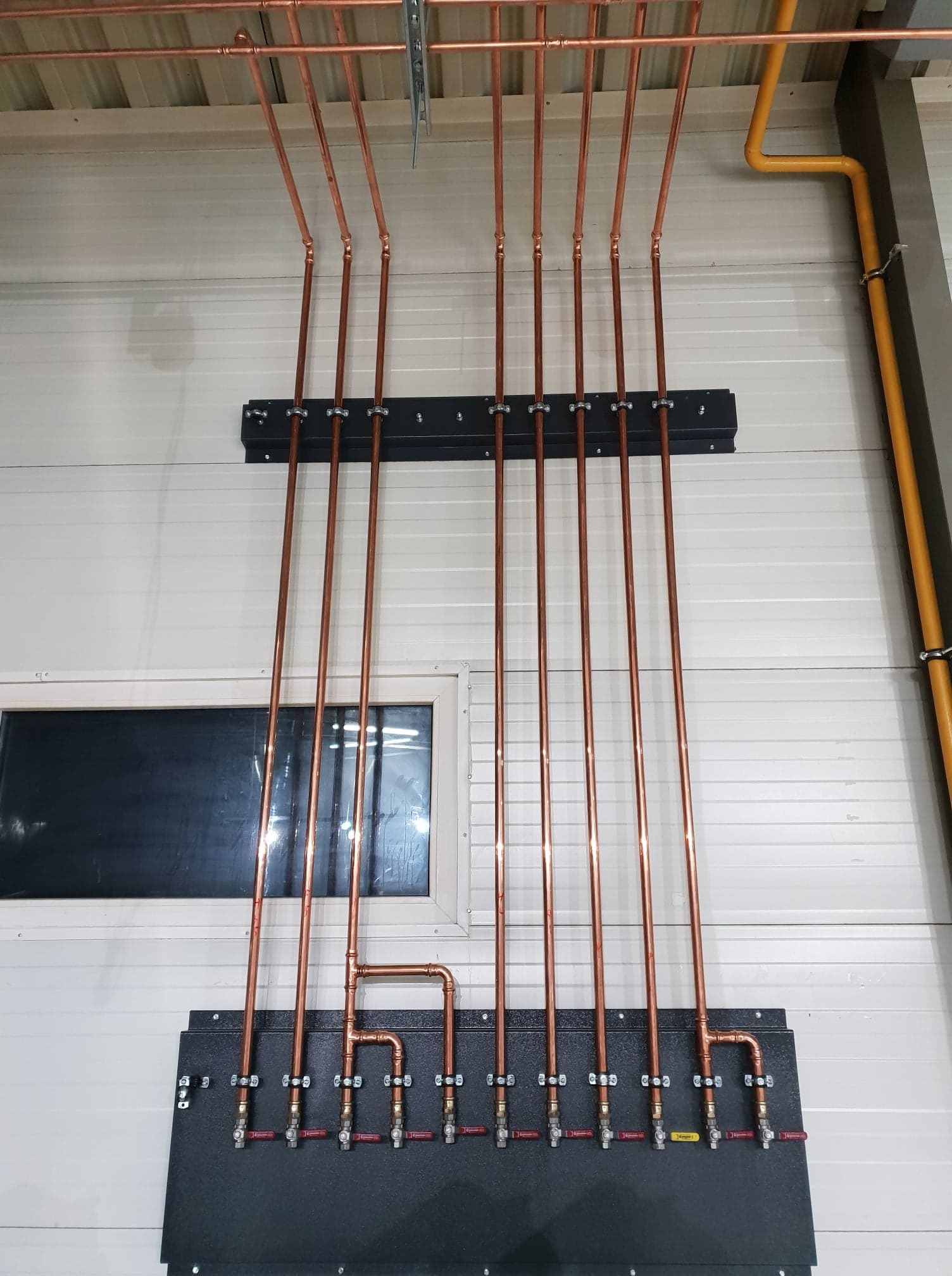 Miedziane rury instalacji gazowej zamontowane na ścianie z blachy trapezowej, z widocznymi zaworami i czarną skrzynką montażową, żółta rura gazowa w tle.