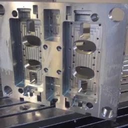 Metalowa forma z licznymi otworami i frezowaniami, leżąca na stole frezarki CNC, widoczne ślady obróbki i chłodziwa.