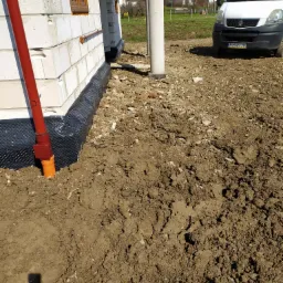 Fundament domu w trakcie prac: widoczna czarna folia izolacyjna przy białych bloczkach, rura spustowa i wykopana ziemia, na tle samochód dostawczy i zabudowa jednorodzinna.