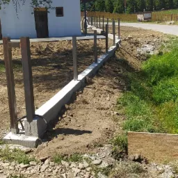 Wylewka betonowa pod ogrodzenie z metalowymi słupkami, widok z perspektywy terenu przed domem jednorodzinnym w słoneczny dzień.