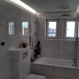 Nowoczesna łazienka wykończona w jasnych, błyszczących płytkach, z minimalistyczną armaturą, grzejnikiem drabinkowym, roślinami w doniczkach i oświetleniem LED podsufitowym.