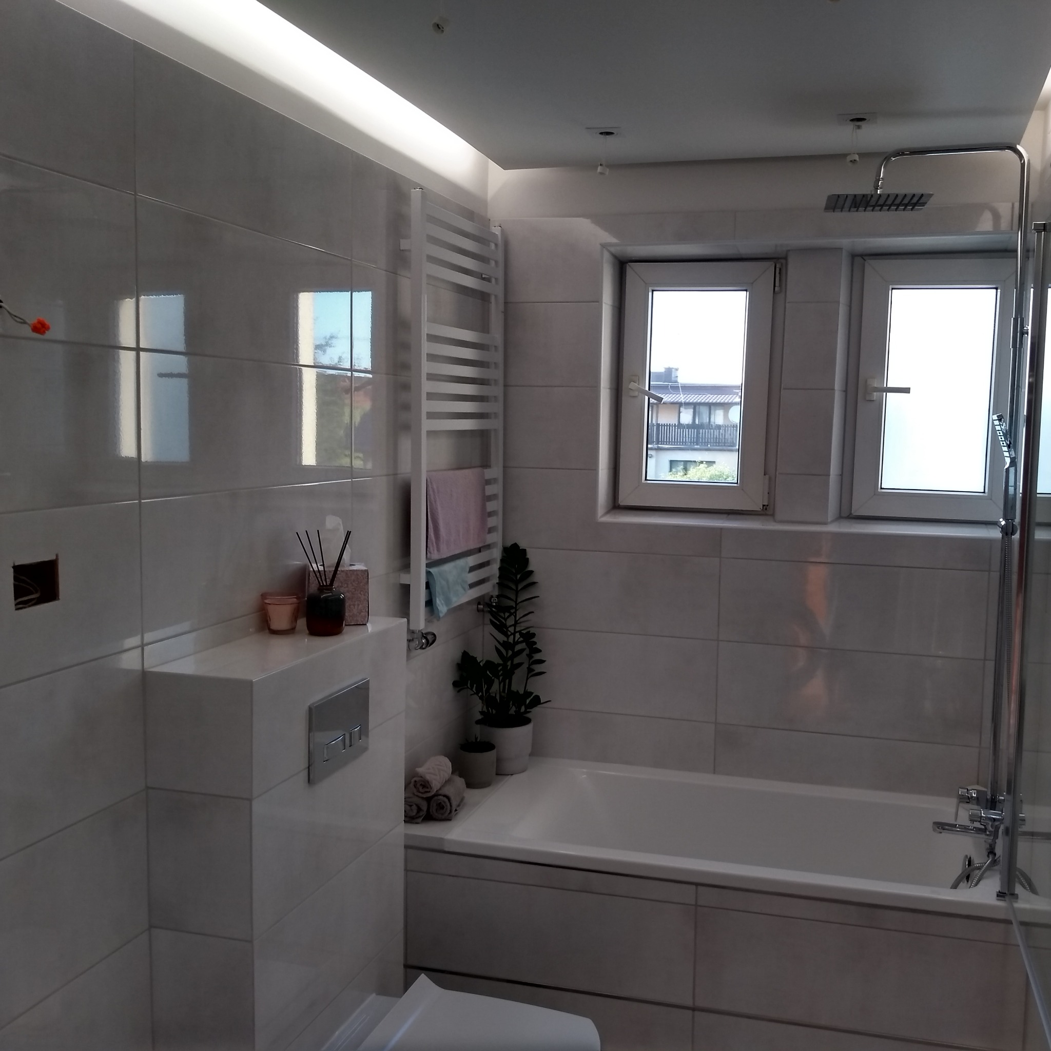 Nowoczesna łazienka wykończona w jasnych, błyszczących płytkach, z minimalistyczną armaturą, grzejnikiem drabinkowym, roślinami w doniczkach i oświetleniem LED podsufitowym.