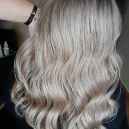 Włosy blond z delikatnymi falami, widok z tyłu, na nadgarstku osoby wykonującej zabieg widoczna czarna bransoletka z napisem LOVE.