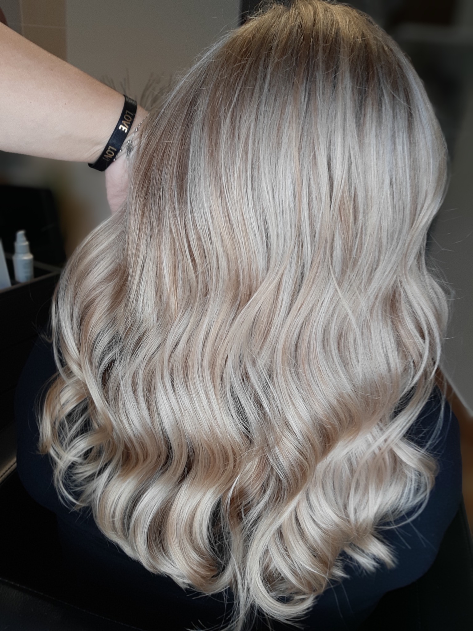 Włosy blond z delikatnymi falami, widok z tyłu, na nadgarstku osoby wykonującej zabieg widoczna czarna bransoletka z napisem LOVE.