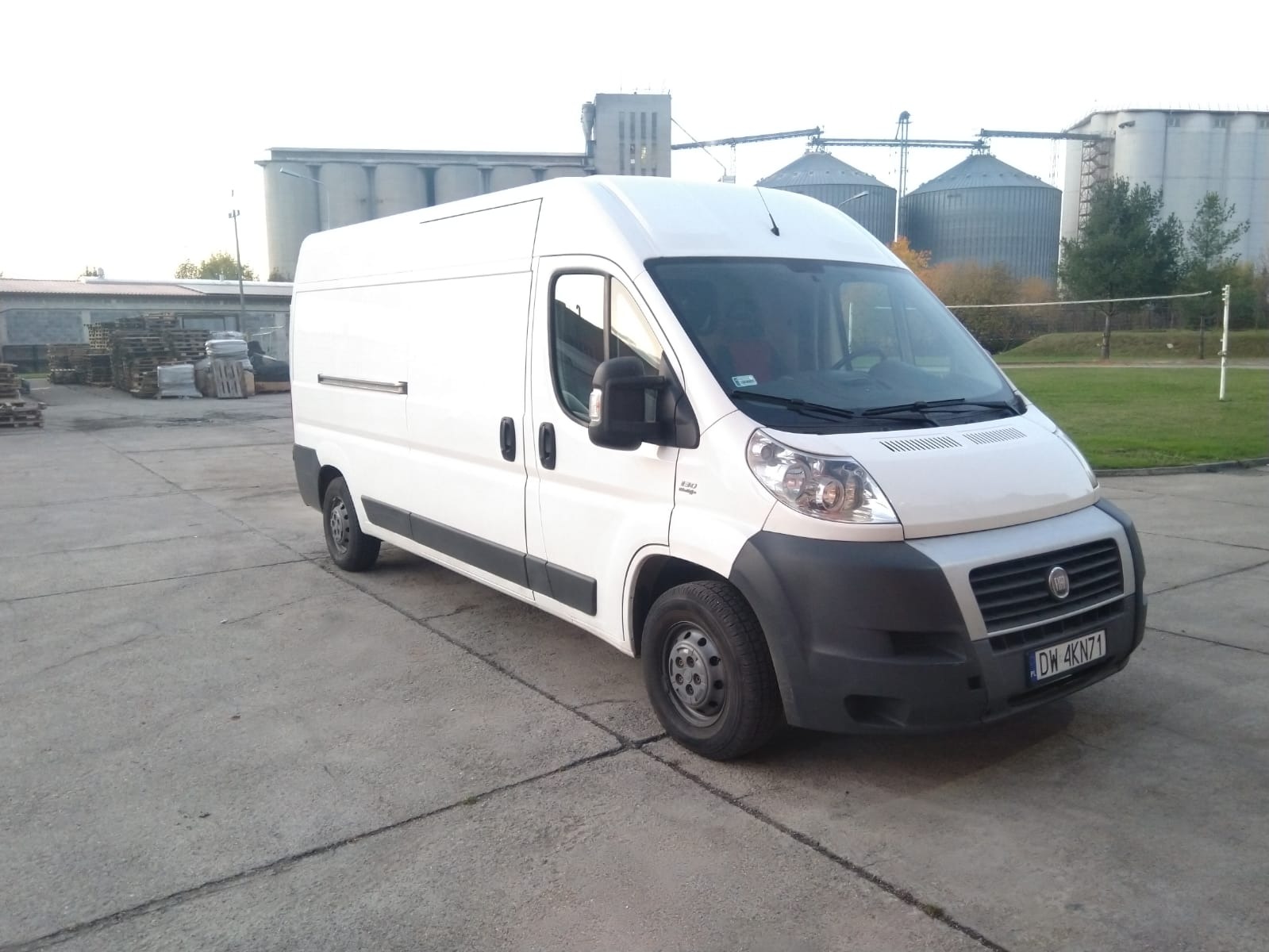 Biały samochód dostawczy Fiat Ducato na betonowym placu, w tle silosy zbożowe i ułożone palety.