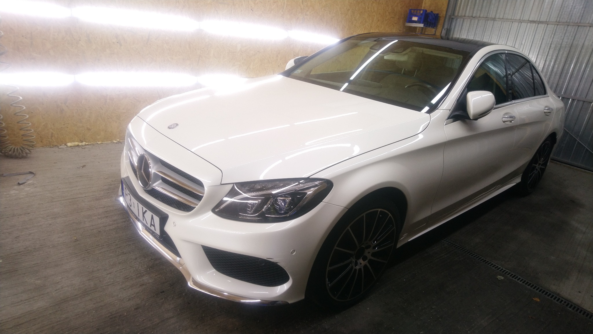 Biały samochód marki Mercedes po umyciu, widoczne krople wody na karoserii, oświetlenie warsztatowe odbija się w lakierze.