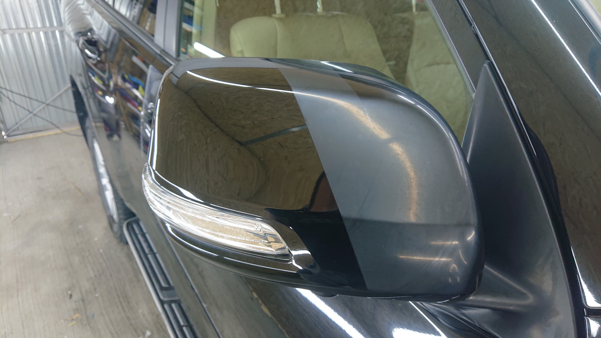 Błyszczące, czarne lusterko boczne samochodu po detailingowym polerowaniu, widoczny wyraźny kontrast między obszarem po zabiegu a obszarem przed, zarys wnętrza pojazdu w tle.