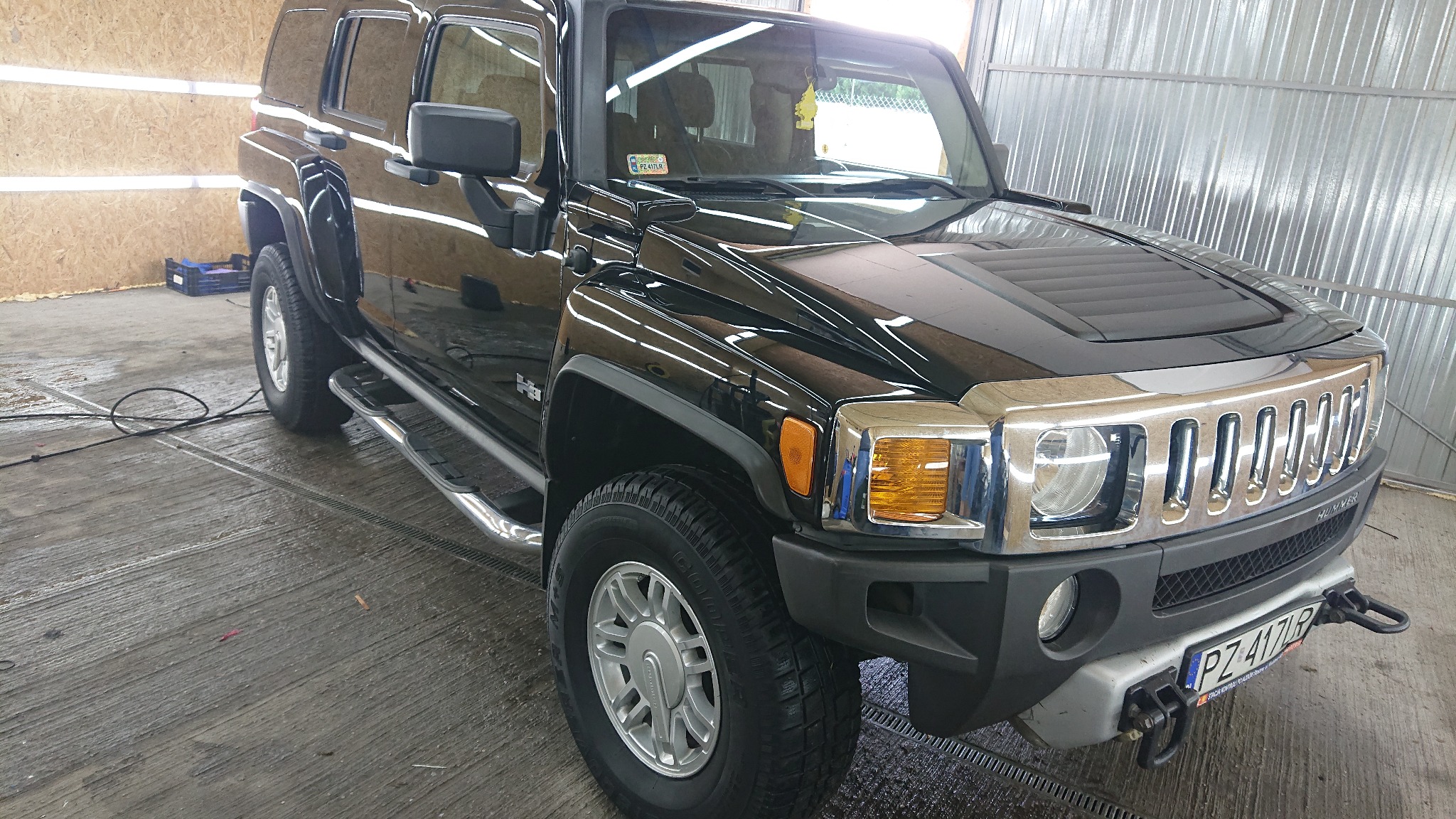 Błyszczący, czarny Hummer H3 po umyciu w myjni, widoczne krople wody na karoserii i mokra posadzka, chromowane elementy grilla i reflektorów, oraz tablica rejestracyjna.