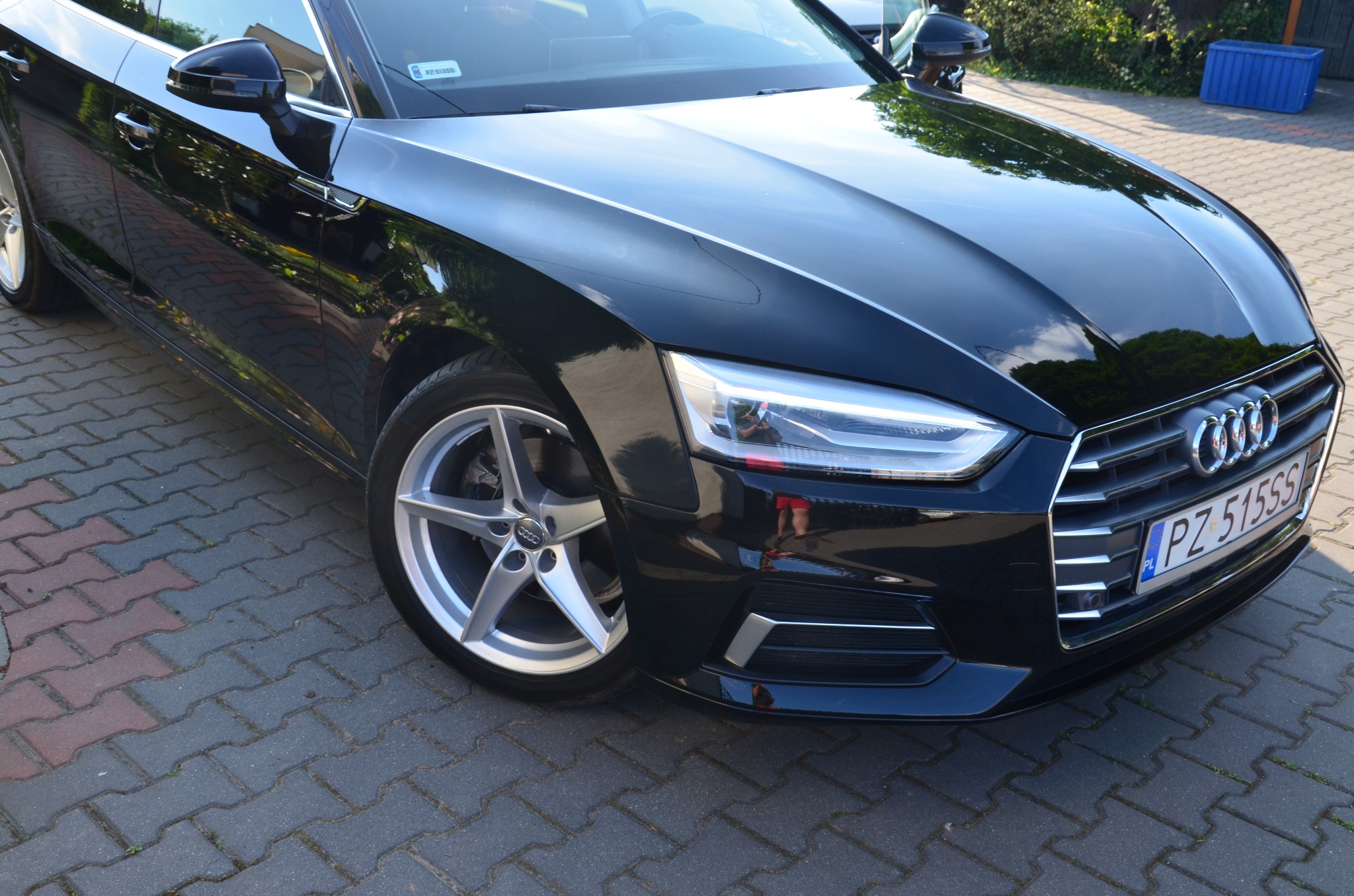 Błyszczący, czarny samochód marki Audi zaparkowany na brukowanym podjeździe, widok z przodu na reflektor, grill i aluminiową felgę.