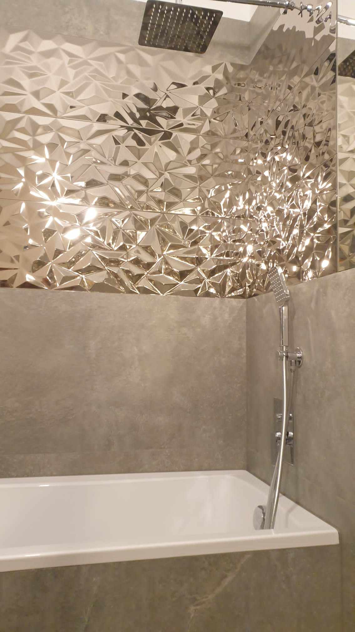 Nowoczesna łazienka z wanną, deszczownicą i ścianą wyłożoną trójwymiarowymi, lustrzanymi płytkami w geometryczny wzór, odbijającymi światło.