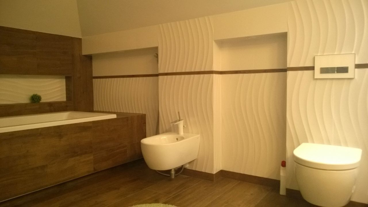 Nowoczesna łazienka z falistą teksturą na ścianach, drewnopodobną zabudową wanny, białym bidetem i toaletą, oraz brązowymi akcentami dekoracyjnymi.