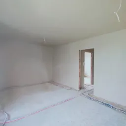 Wnętrze surowego pomieszczenia po tynkowaniu, widoczne wystające przewody instalacji elektrycznej i rury ogrzewania podłogowego, ściany i sufit pokryte świeżym, jasnoszarym tynkiem, surowy stan...