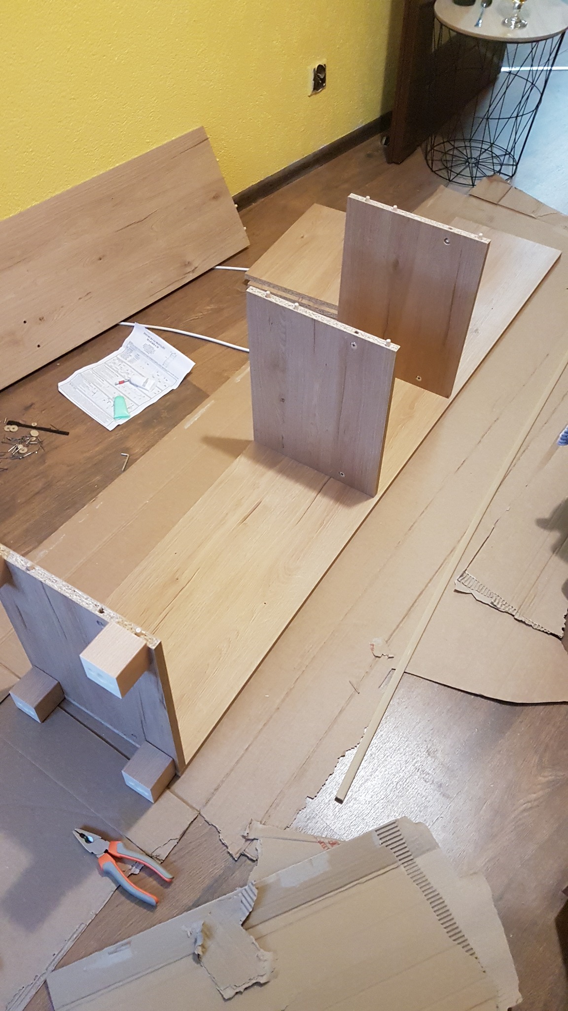 Składanie mebli z drewnianych płyt na podłodze zabezpieczonej kartonem, widoczne narzędzia i instrukcja montażu.