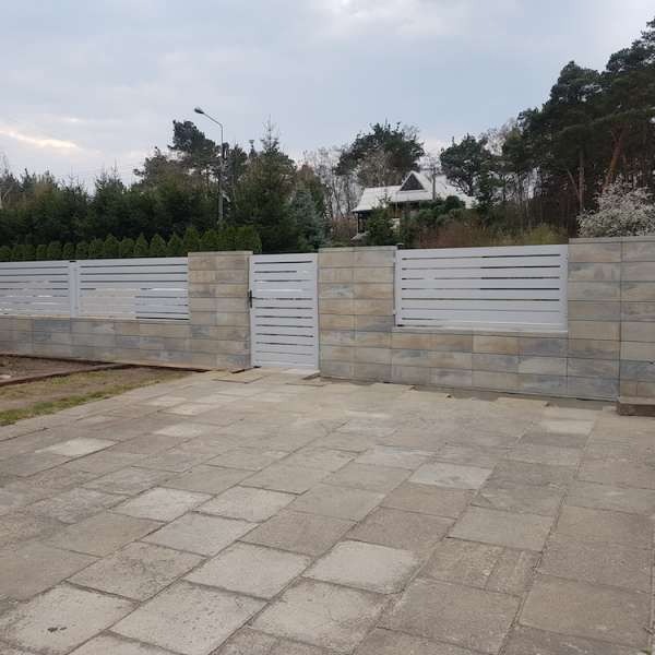 Nowoczesne ogrodzenie z białymi, poziomymi przęsłami i szarą podmurówką z bloczków betonowych, brama furtka wkomponowana w ogrodzenie, widok z kostki brukowej na tle domu i drzew