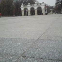 Rozległy plac wyłożony szarymi płytami, w tle arkady i drzewa bez liści, pochmurny dzień.