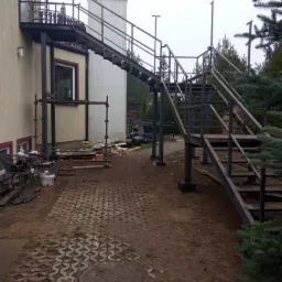 Zewnętrzne, dwubiegowe schody z metalu z platformą, prowadzące do budynku, widoczne elementy konstrukcyjne i niedokończone wykończenie terenu.