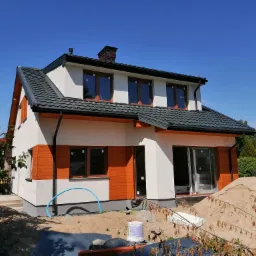 Dwukondygnacyjny dom z fragmentami elewacji w kolorze białym i pomarańczowym, ciemna dachówka, widoczny fragment ogrodu i sterta piasku przed domem.