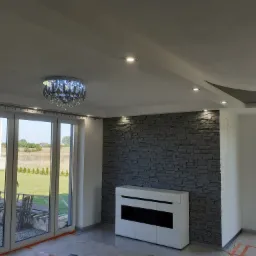 Wnętrze pokoju z dekoracyjną ścianą z szarej cegły, białym meblem RTV, dużym oknem z widokiem na zieleń i ozdobnym żyrandolem kryształowym.