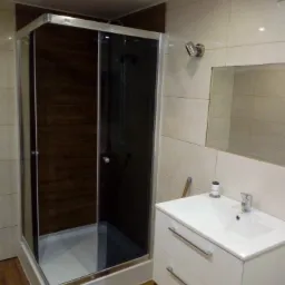 Nowoczesna kabina prysznicowa z ciemnymi płytkami imitującymi drewno, umywalka z szafką w jasnym odcieniu, oświetlenie punktowe i lustro na ścianie wyłożonej beżowymi płytkami.