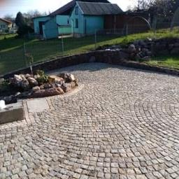 Perfekcyjne promocyjne kompleksowe usługi brukarskie - Ułożona z granitowej kostki podjazd w kształcie wachlarza, obrzeże z kostki, w tle dom i skarpa z kamieniami.
