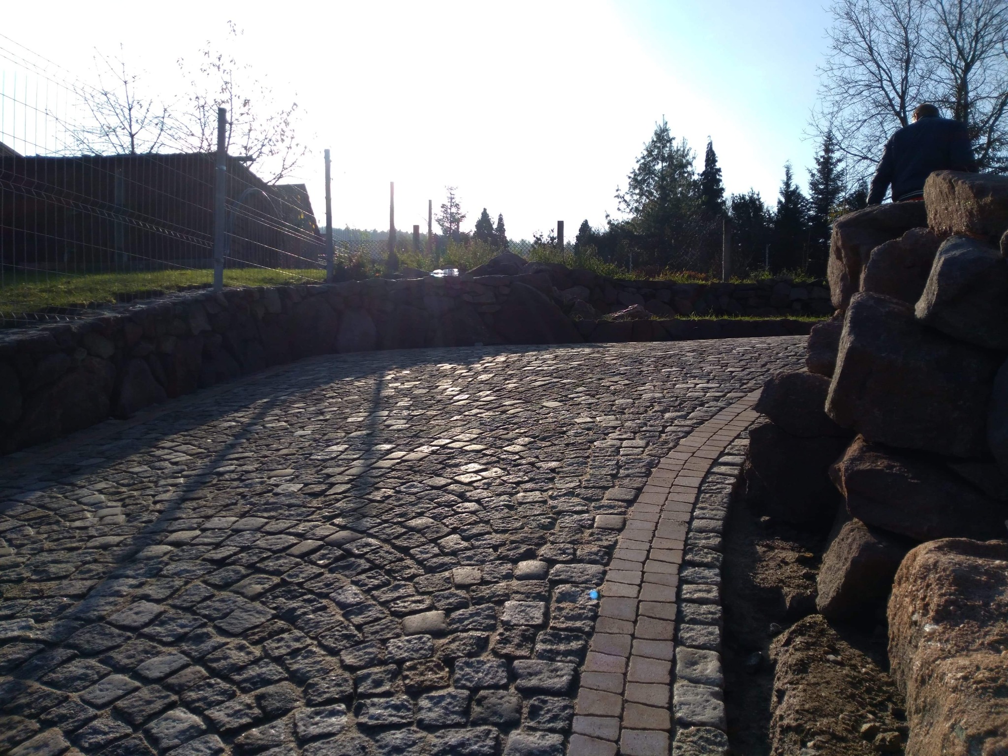 Ułożona z kostki granitowej ścieżka w ogrodzie, obrzeża z jasnej kostki, w tle murki z kamienia i zieleń, po prawej stronie ułożone duże kamienie, na których stoi osoba.