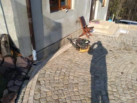 Układanie nawierzchni z kostki granitowej o łukowatym wzorze przy domu, widoczna zagęszczarka i cień osoby robiącej zdjęcie.