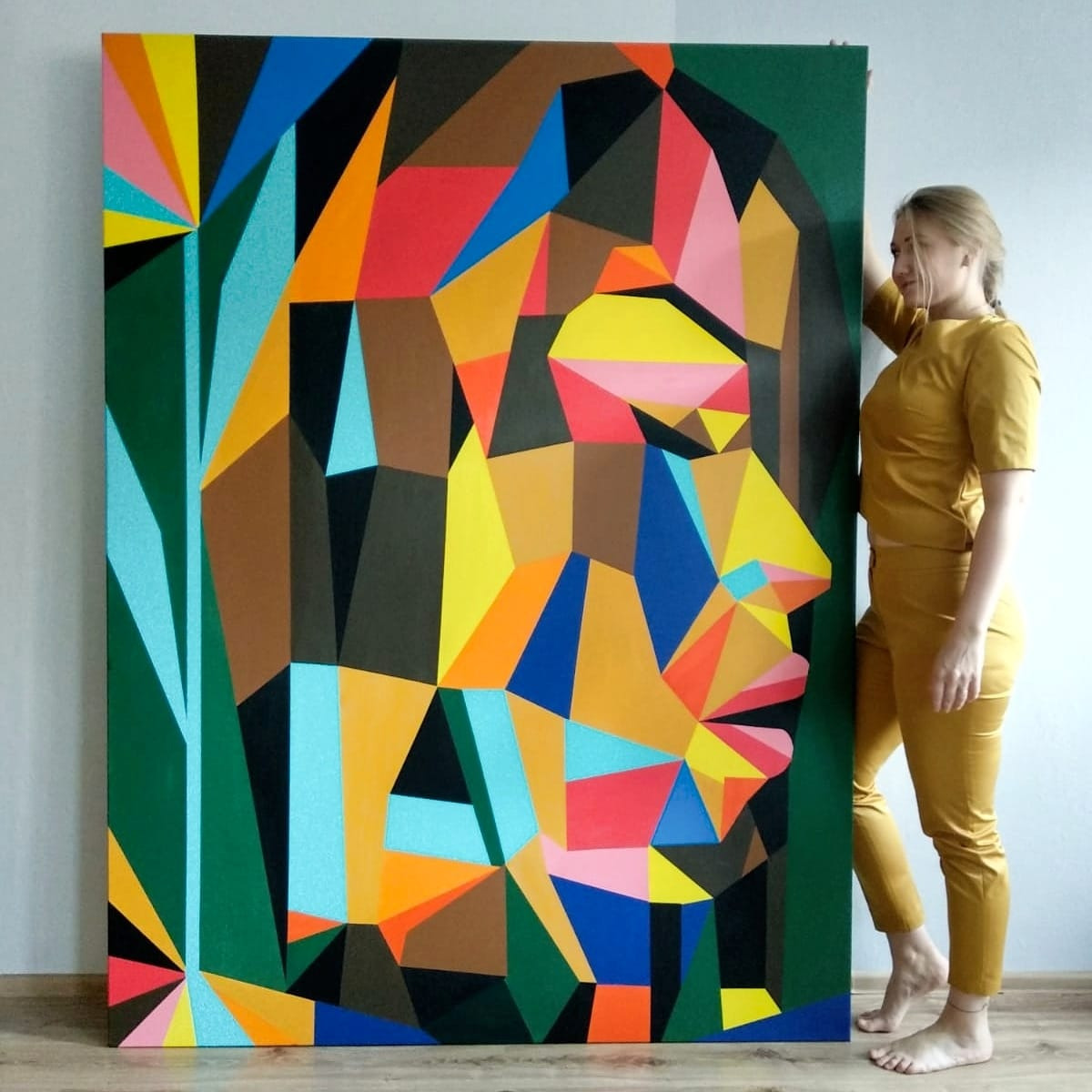 Kobieta w żółtym stroju opiera się o duże, geometryczne malowidło portretowe w odcieniach brązu, zieleni, żółci, czerwieni i niebieskiego, z wyraźną fakturą farby.