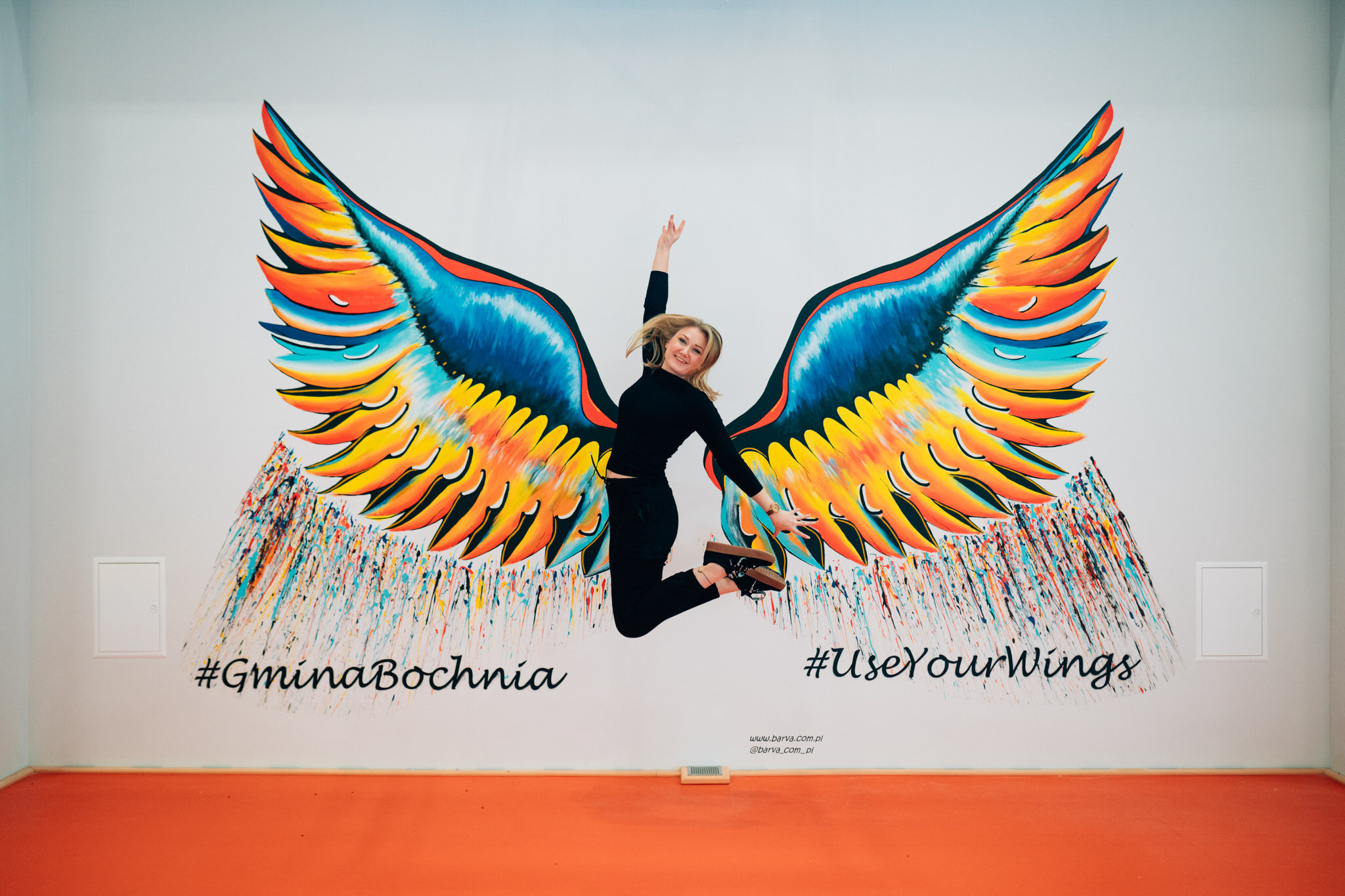Kobieta skacze przed muralem ze skrzydłami anioła we Wrocławiu, z hasłami #GminaBochnia i #UseYourWings.