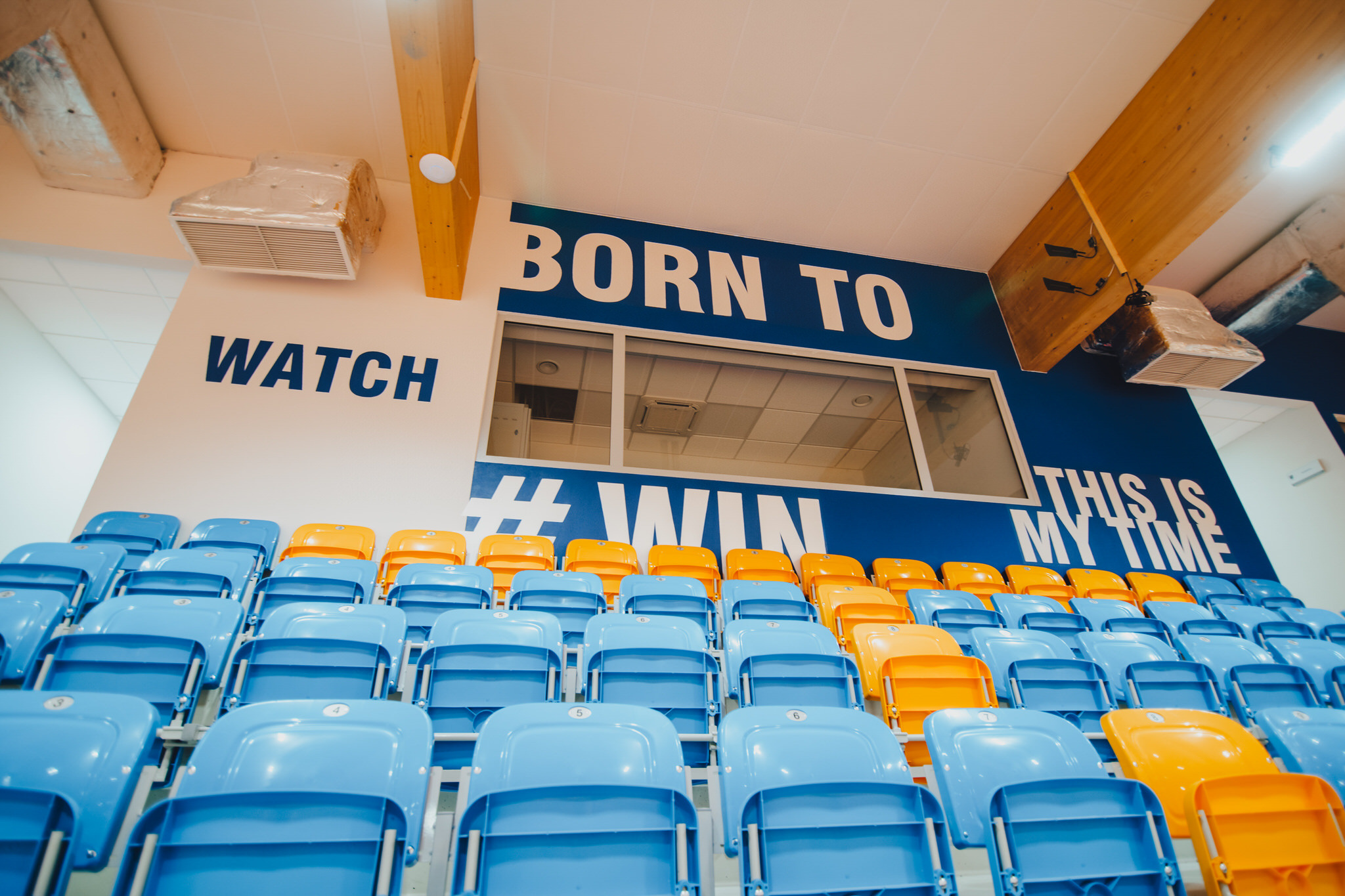 Widok z dołu na trybuny stadionu z niebieskimi i pomarańczowymi krzesełkami, na ścianie napis 'WATCH BORN TO #WIN THIS IS MY TIME', widoczne elementy konstrukcyjne pod sufitem.