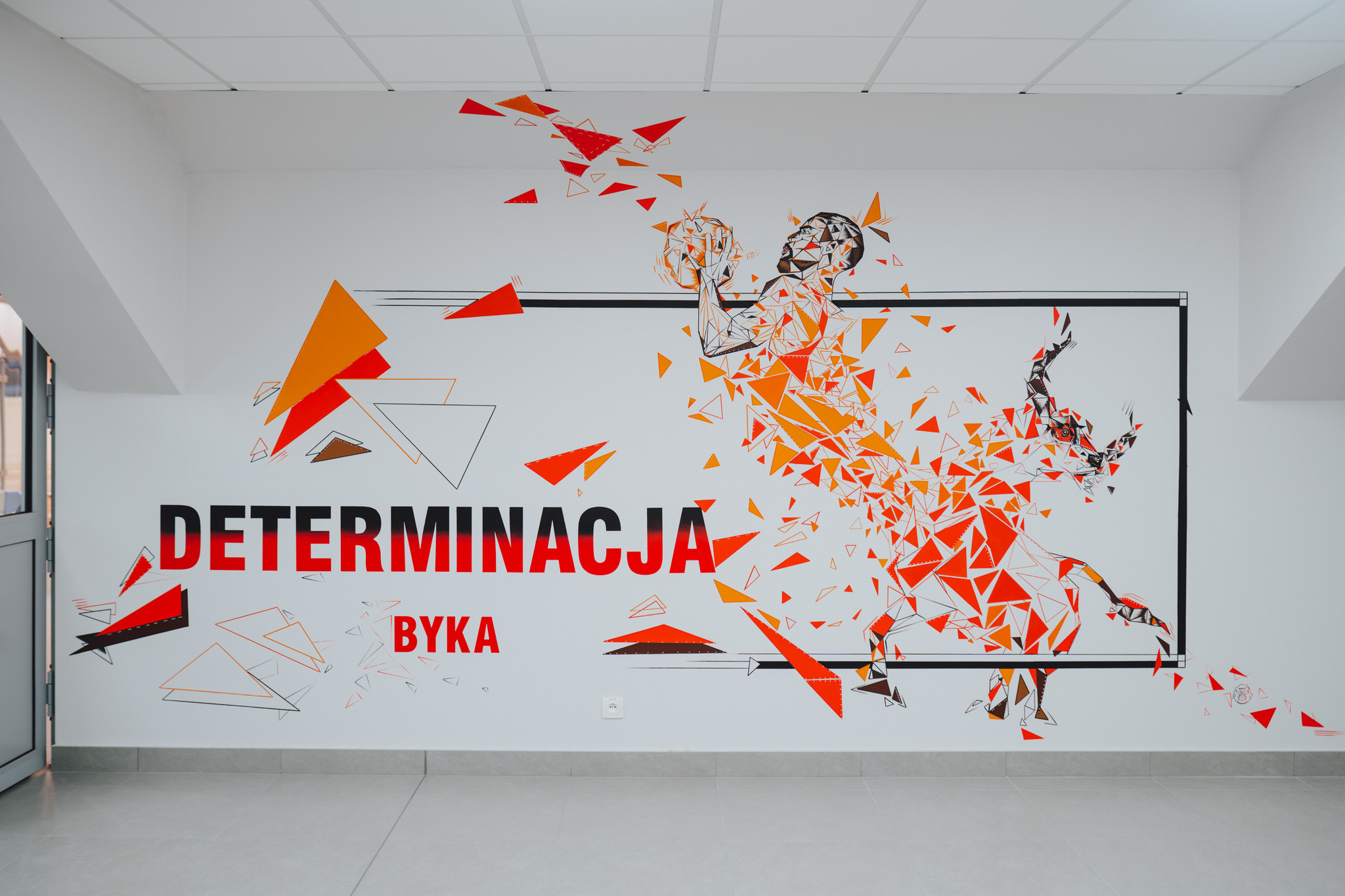 Grafika ścienna przedstawiająca koszykarza w dynamicznej pozie, wykonana z czerwono-pomarańczowych trójkątów, z napisem 'DETERMINACJA BYKA' w czerni i czerwieni.