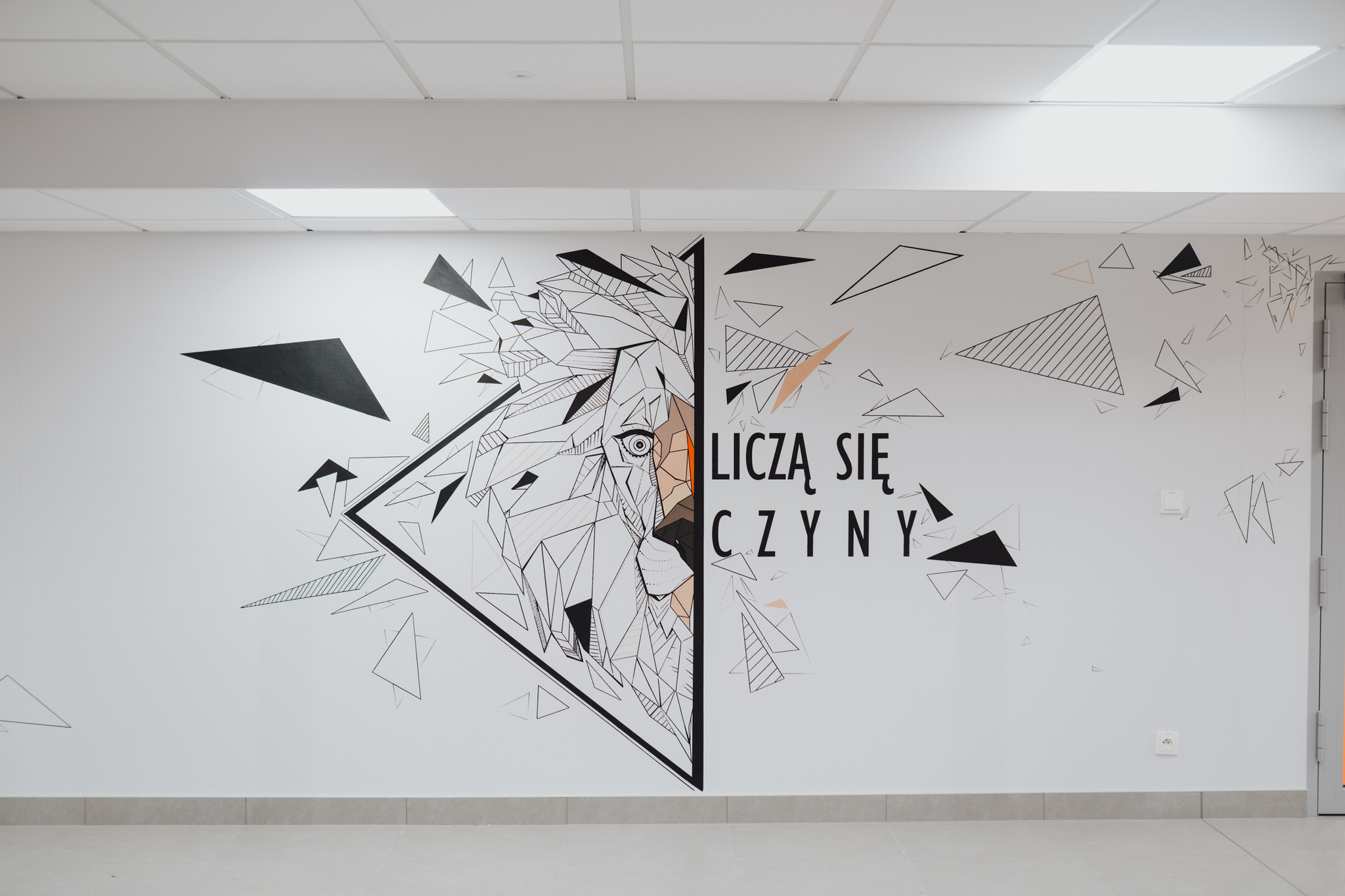 Geometryczny mural lwa z napisem 'Liczą się czyny' na białej ścianie w biurze, widok z dołu.