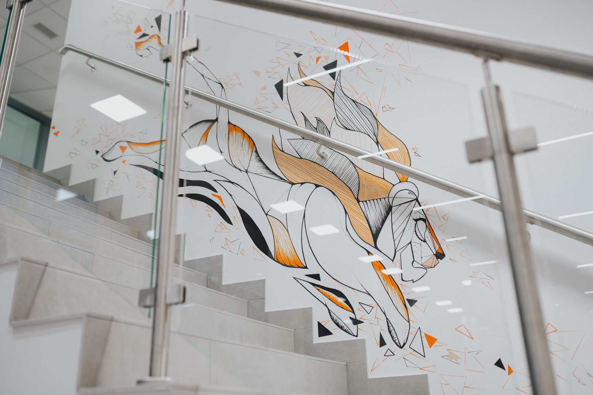 Ilustracja lwa w stylu geometrycznym, wykonana czarnymi, pomarańczowymi i białymi liniami na białej ścianie przy schodach z szarymi stopniami i szklaną balustradą.