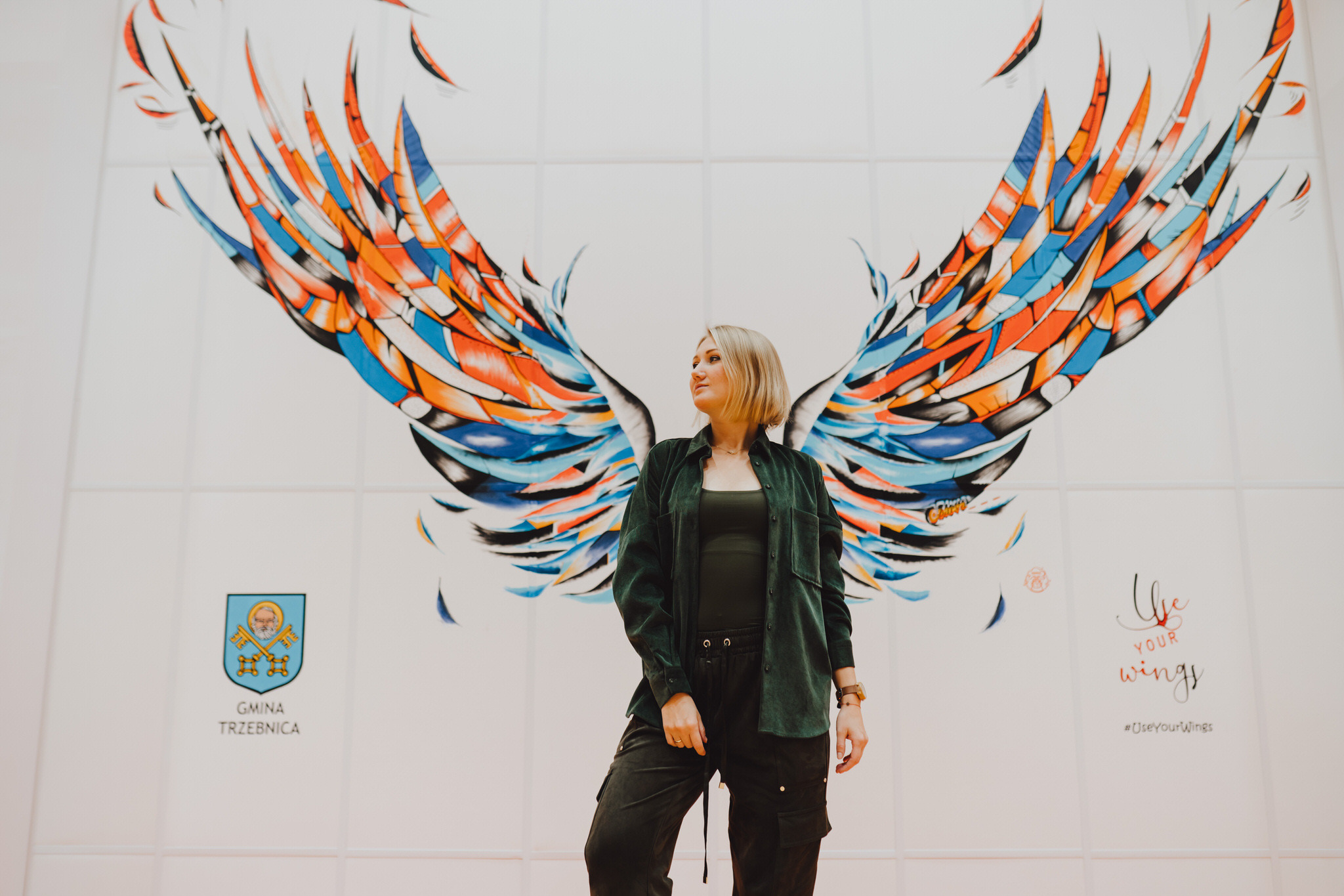 Kobieta pozuje na tle muralu przedstawiającego kolorowe skrzydła anioła na ścianie z logo Gminy Trzebnica i napisem 'Use your wings'.