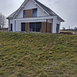 Nowoczesny dom z białą elewacją i drewnianymi panelami dekoracyjnymi, widoczny z perspektywy trawnika w pochmurny dzień.