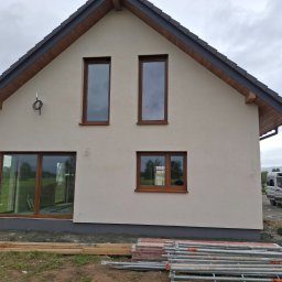 ŁUKASZ KOWNACKI - Elewacja domu jednorodzinnego z brązowymi oknami, widoczne rusztowania i materiały budowlane. Świeżo otynkowana ściana w jasnym odcieniu i ciemny dach.