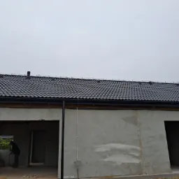 Szary budynek w trakcie budowy z nowym, ciemnym dachem z dachówki ceramicznej. Widoczne niezabezpieczone otwory okienne i drzwiowe. Na pierwszym planie elewacja.