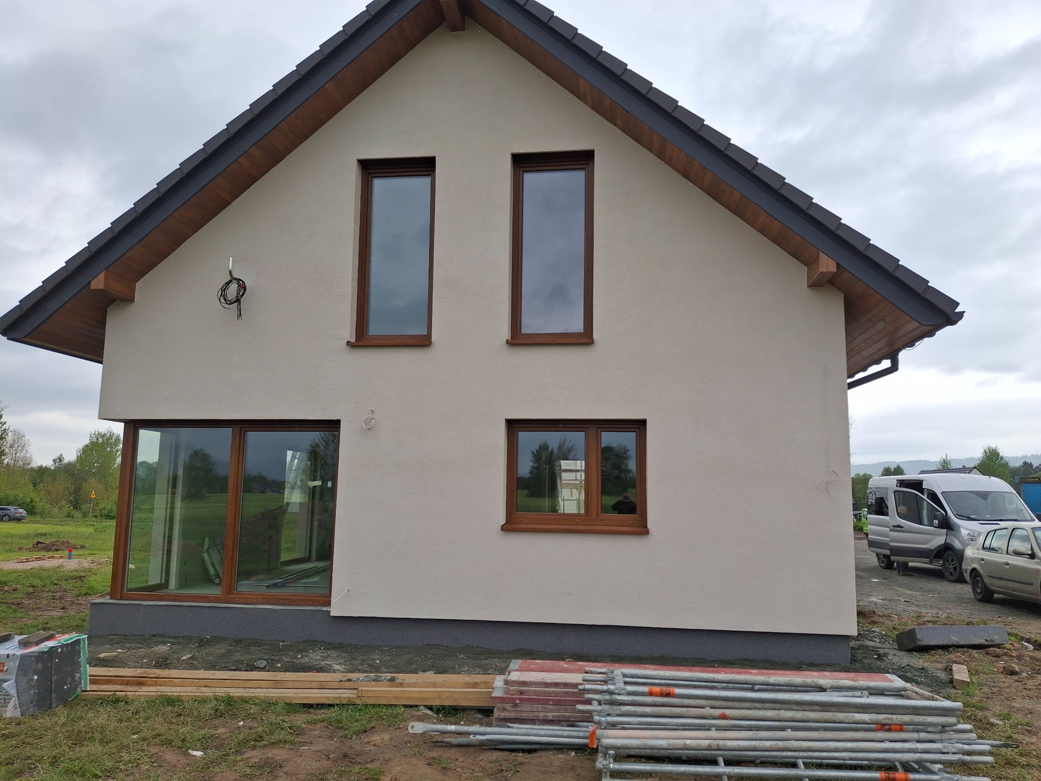 Elewacja domu jednorodzinnego z brązowymi oknami, widoczne rusztowania i materiały budowlane. Świeżo otynkowana ściana w jasnym odcieniu i ciemny dach.