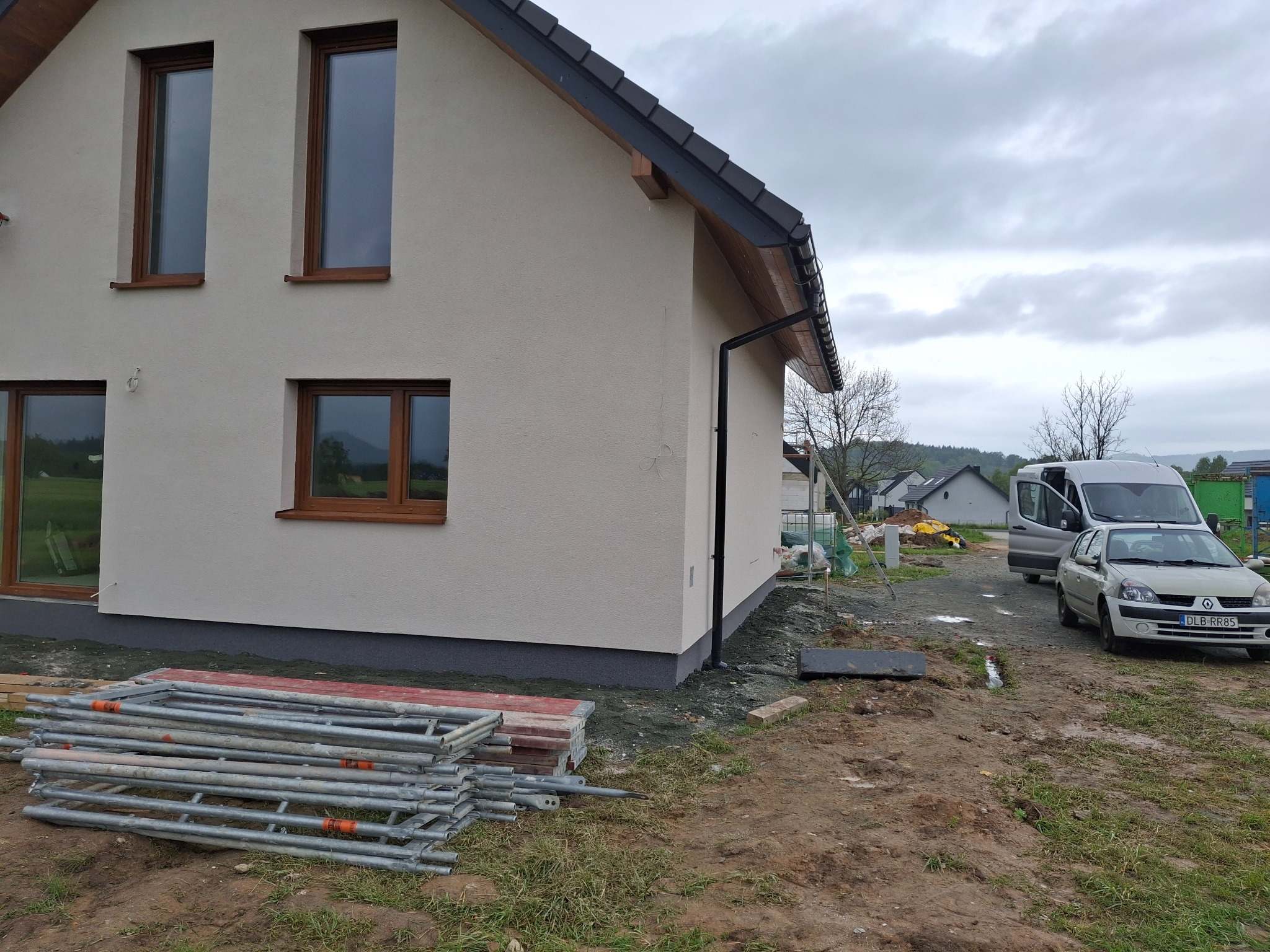 Nowoczesny, kremowy dom jednorodzinny z brązowymi oknami i ciemnym dachem. Na pierwszym planie rusztowanie i teren budowy, w tle zaparkowane samochody.