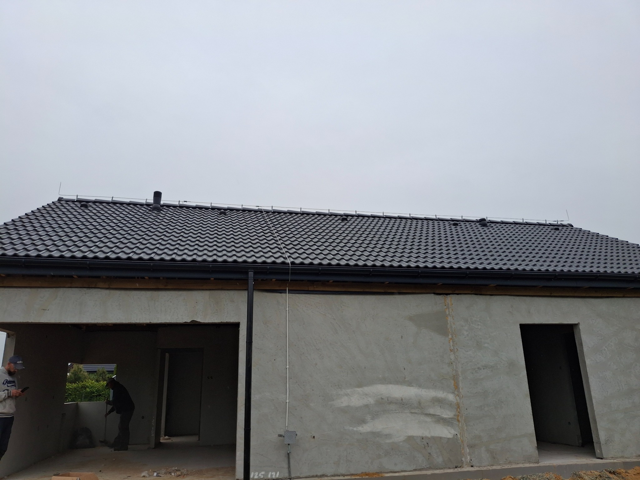 Szary budynek w trakcie budowy z nowym, ciemnym dachem z dachówki ceramicznej. Widoczne niezabezpieczone otwory okienne i drzwiowe. Na pierwszym planie elewacja.