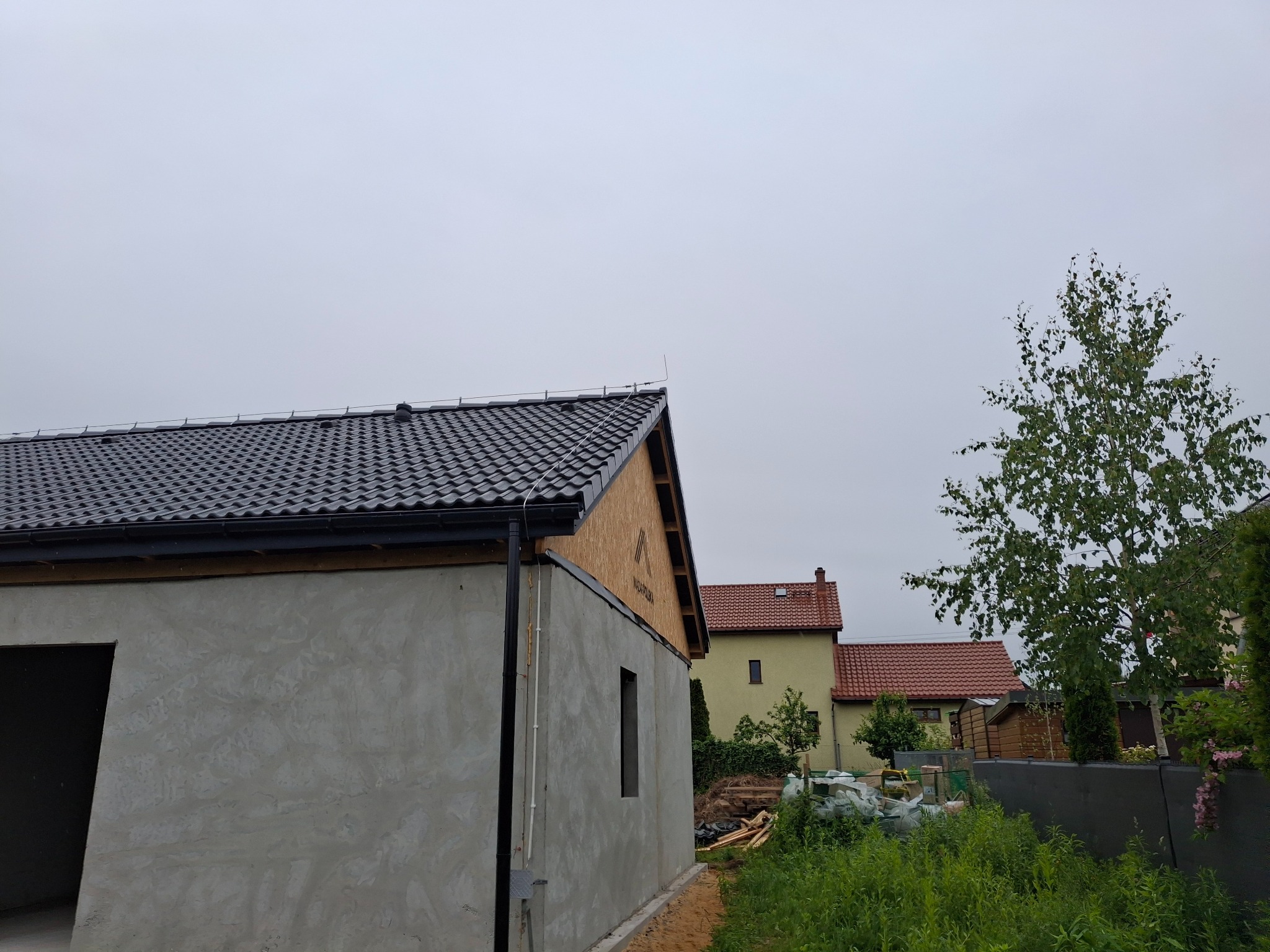 Surowy budynek z ciemną dachówką, widoczne tynki i fragment elewacji z desek. W tle zabudowa jednorodzinna i zieleń w pochmurny dzień.