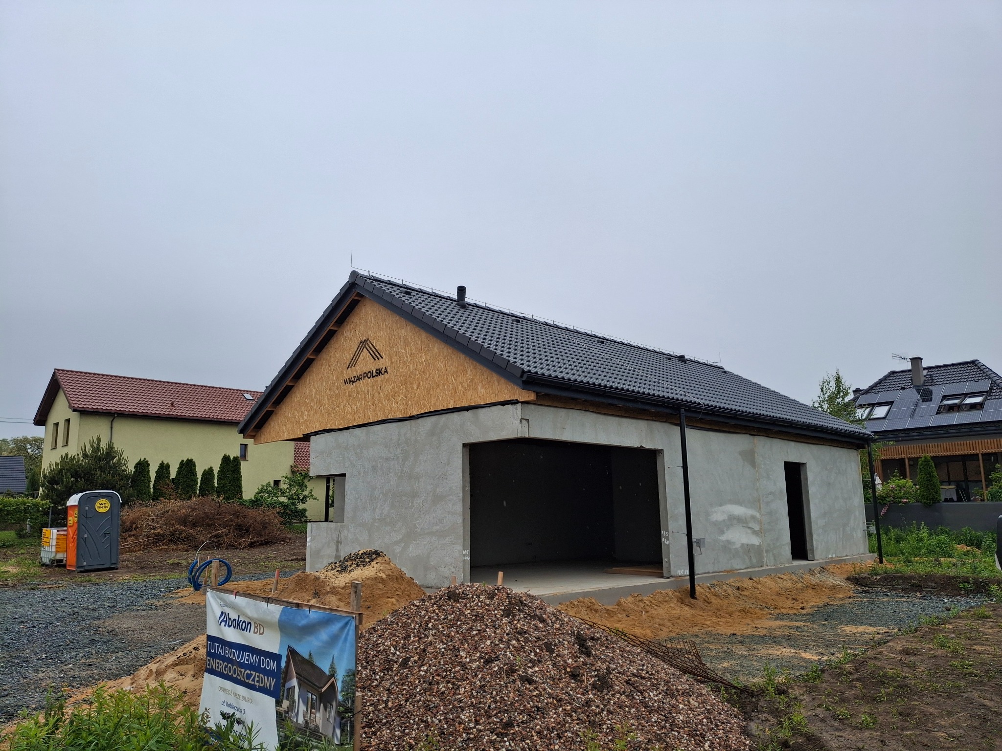 Surowy budynek gospodarczy z dachem pokrytym czarną dachówką, w trakcie budowy, widoczna płyta OSB z logo firmy. Na pierwszym planie hałda żwiru i baner reklamowy.