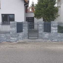 Nowoczesne ogrodzenie z szarych bloczków betonowych i metalowych przęseł, z bramą wejściową, na tle domu z drewnianymi zdobieniami okiennymi.