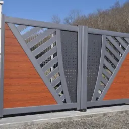 Nowoczesna brama wjazdowa w Bolesławcu z drewnianymi i metalowymi elementami, o geometrycznym wzorze, osadzona na kamiennych słupkach.