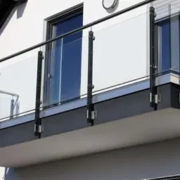Nowoczesny balkon z szklaną balustradą i czarnymi elementami mocującymi, widok z dołu, jasna elewacja budynku w tle.