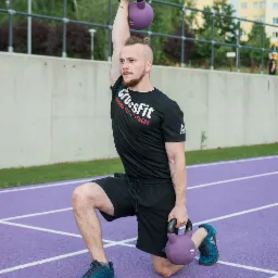 Mężczyzna w czarnej koszulce CrossFit wykonuje wykrok z dwoma fioletowymi kettlebells na fioletowej bieżni.